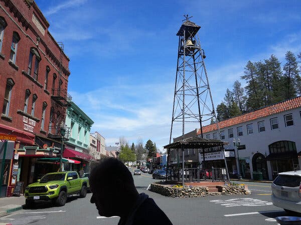 A silhouetted head is shown in the foreground. Behind it is a view of a street in a small town. In the street, a bell tower rises. 