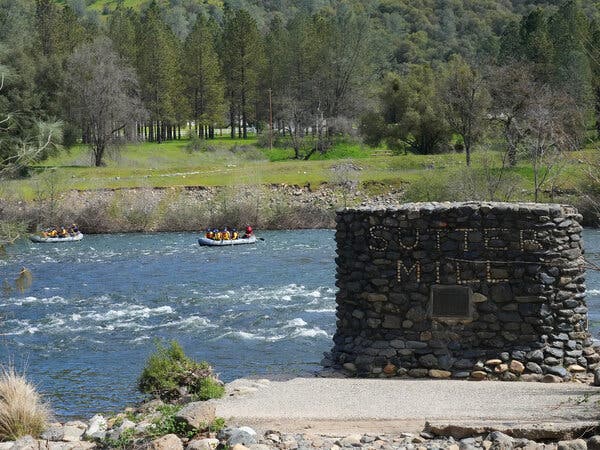 Groups of rafters ride down a river in a park. In the foreground sits a stone structure